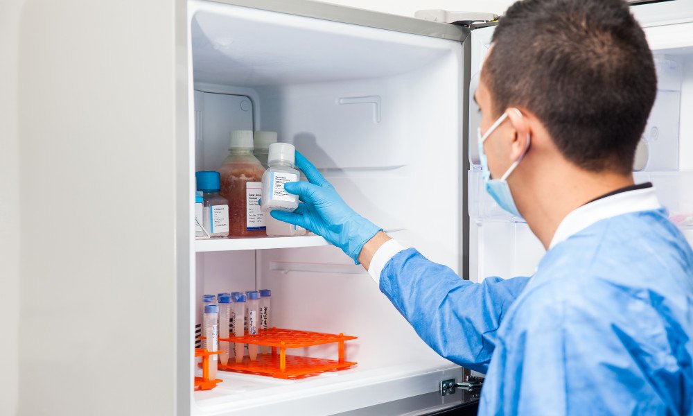 Profissional de laboratório, vestindo jaleco e luva cirúrgica azuis claros, verificando a validade dos insumos laboratoriais em uma geladeira.